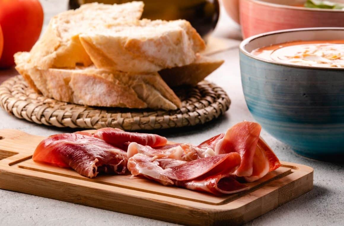 Pe un tocător de bucatarie din plemn sunt asezate mai multe felii de jamon serrano, iar alaturi sunt cateva felii de paine si un bol ceramic cu supă. Diferența între jamon și prosciutto
