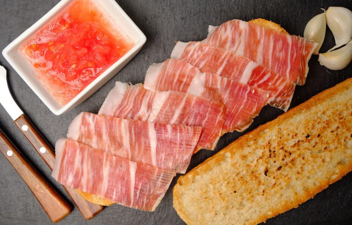 mai multe felii de prosciutto sunt asezate pe o felie mare de paine prajita, iar alaturi de ea mai e o felie de paine prajita si un recipient alb cu sos de rosii, două tacâmuri și trei cătei de usturoi