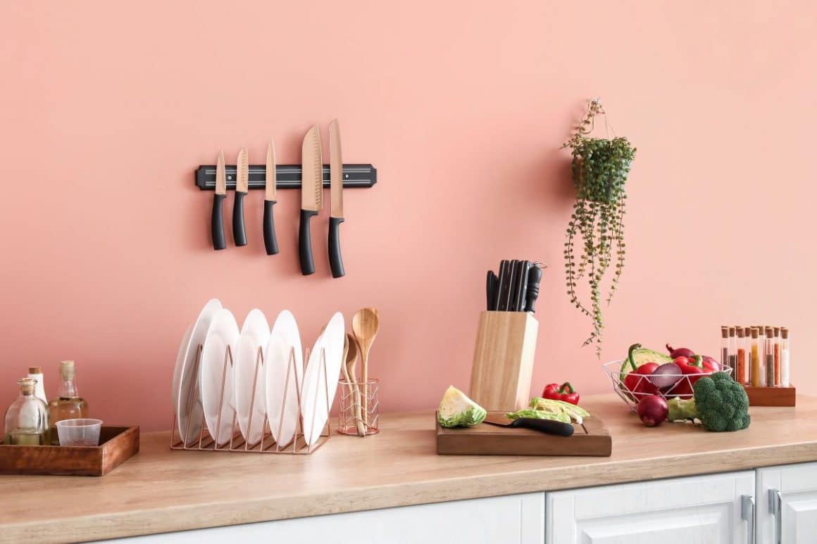 mai multe cutite, ustensile esențiale într-o bucătăriesunt dispuse pe un suport de lemn prins pe un perete gri, alaturi de mai multe ingrediente naturale și sticle