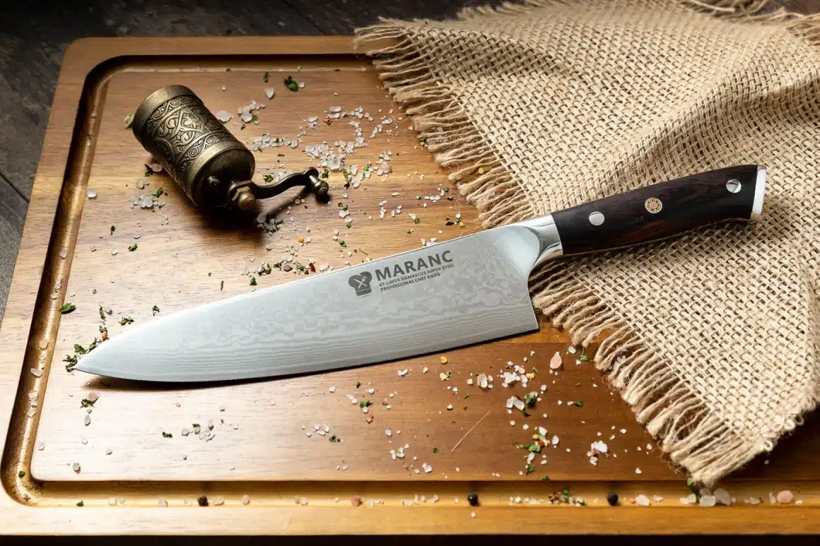 un cutit de bucatarie este asezat pe un tocător, cu o panza naturala langa el și mai multe cristale de sare și o rasnita de alama