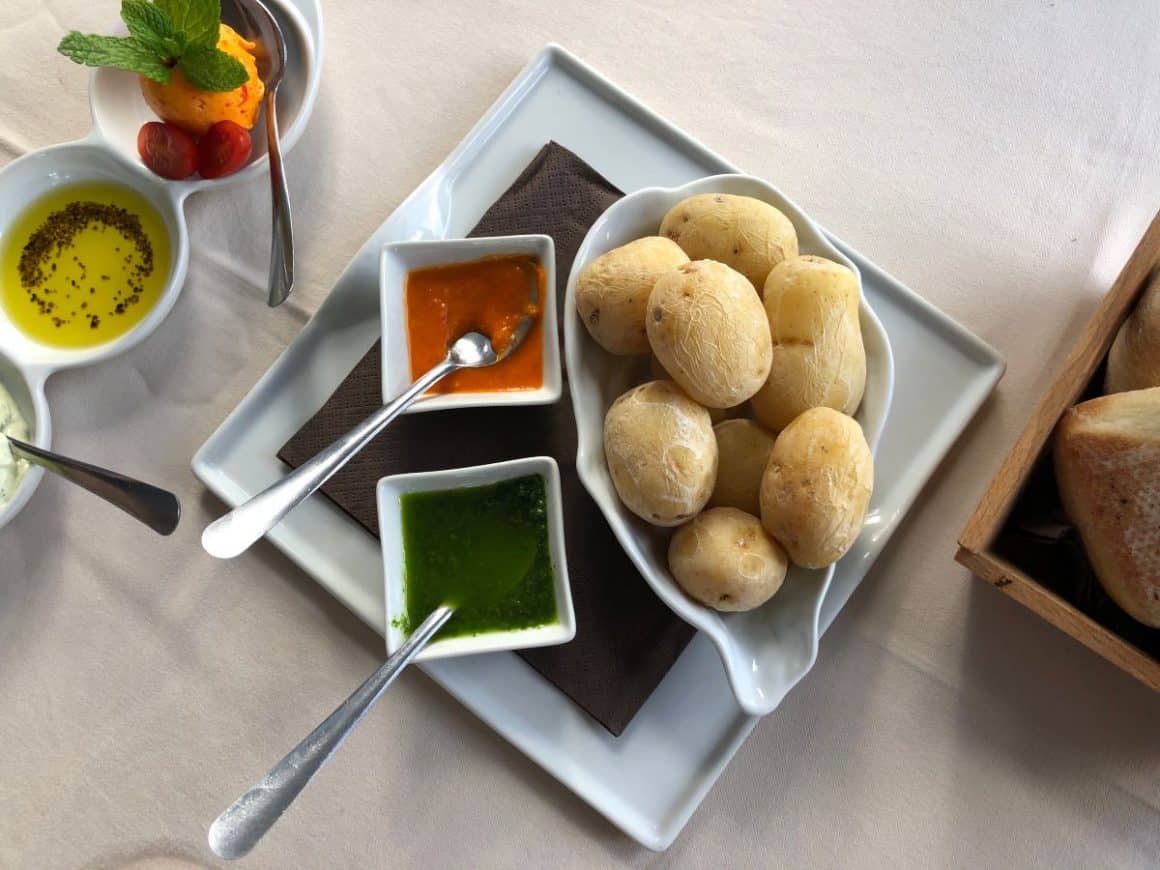 o portie de Papas arrugadas cu cartofi fierti și doua recipiente cu sos mojo rosu și sos mojo verde sunt asezate alaturi de tacamuri pe o masa alba