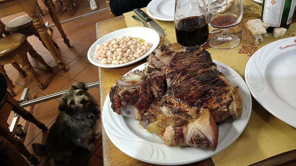 o portie de Bistecca alla fiorentina, una din mancarurile traditionale din toscana, este servita pe o masa de lemn, alaturi de o farfurie cu fasole, iar un caine sta sub masă