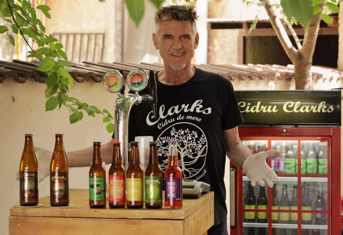 propietarul cidrariei clarks pozeaza intr-o gradina in fata standului cu cidru pe care sunt asezate 7 sticle, iar in spatele lui este un frigider cu sticle de cidru rece