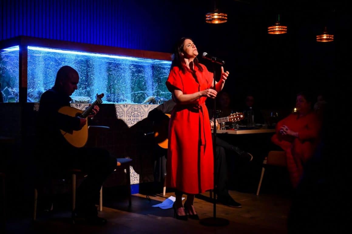 o femeie imbracata intr-o roche rosie canta la microfon pe o scena, iar in spatele ei sunt cativa instumentisti