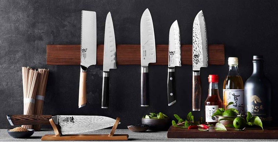 mai multe cutite, ustensile esențiale într-o bucătăriesunt dispuse pe un suport de lemn prins pe un perete gri, alaturi de mai multe ingrediente naturale și sticle