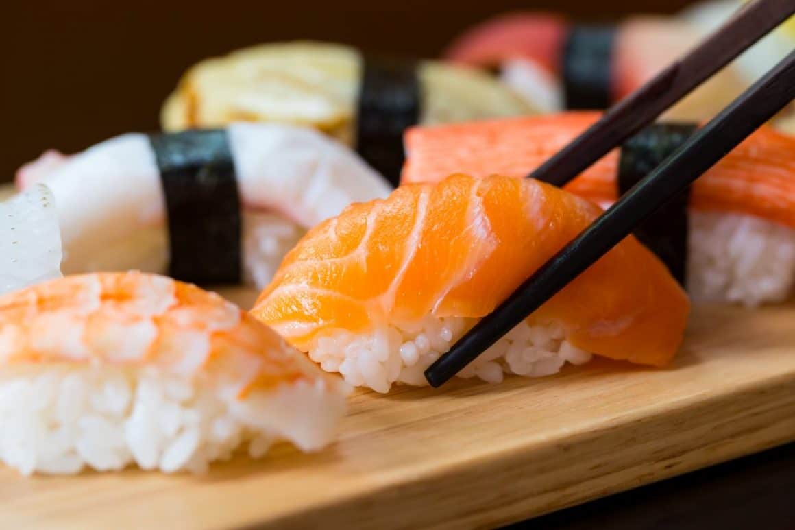 doua betisoare chinezesti apuca o bucata de sushi nigiri de pe unplatou de lemn. Cum se mănâncă sushi