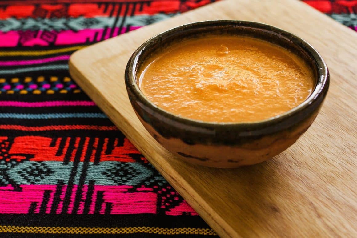 Bol din lut plin cu sosul Habanero, asezat pe un tocator din lemn, pe un stergar cu motive mexicane