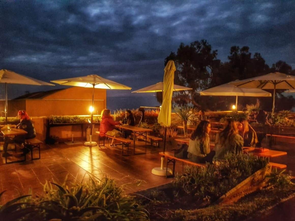 mai multe grupri de femei iau masa pe terasa restaurantului Tacoa printre ghivece cu plante si umbrele, la ora apusului