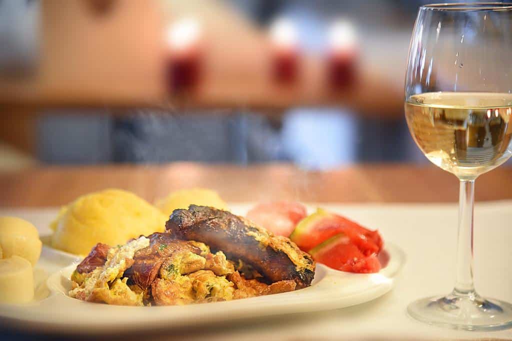 o porție de mancare cu carne și mamaligă sunt asezate pe o farfurie, langa un pahar de vin alb