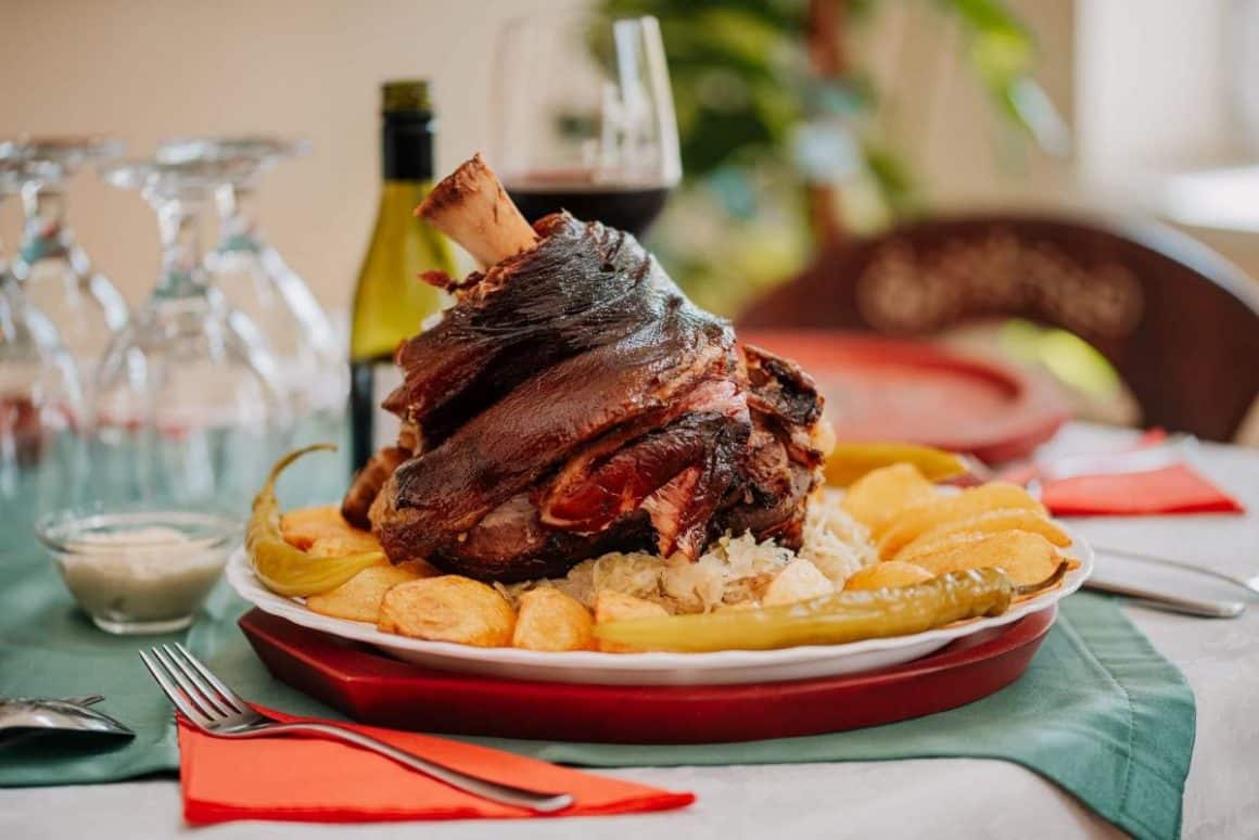 un ciolan de porc rumenit este asezat pe o farfurie, pe un pat de cartofi mici, copți