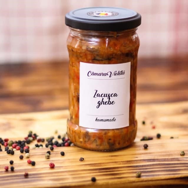 un borcan cu zacusca este asezat pe un tocător de lemn cu mai multe boabe de piper mozaic