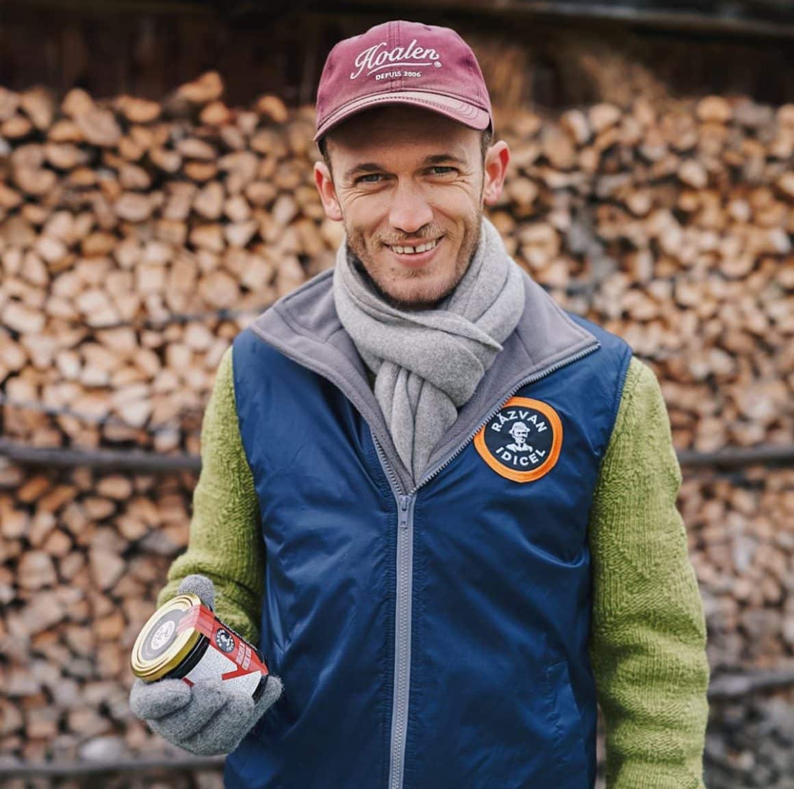 un barbat cu sapcă și vestă zâmbește în timp ce ține în mână un borcan de zacusca. in fundal este o stivă de lemne de foc