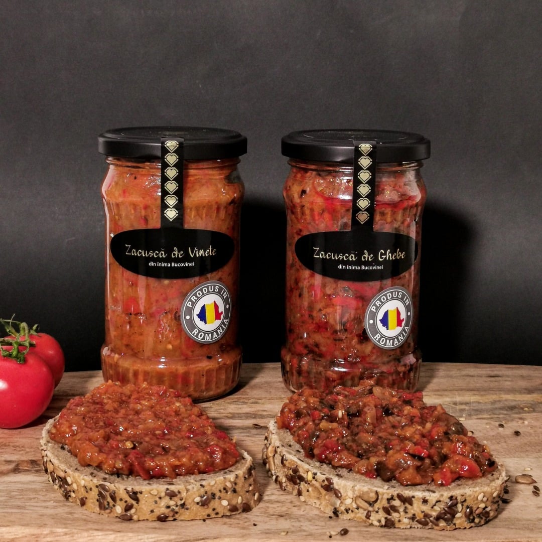 două borcane de zacuscă cu capac negru sunt asezate în fața a două felii de pâine țărănească pline de zacuscă de la producătorii locali