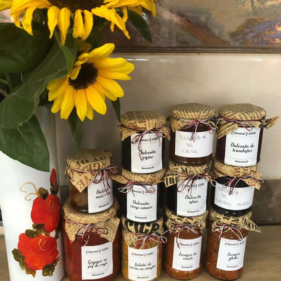 mai multe borcane cu zacusca de la producători locali sunt asezate unu peste altul, langa un buchet de floarea soarelui