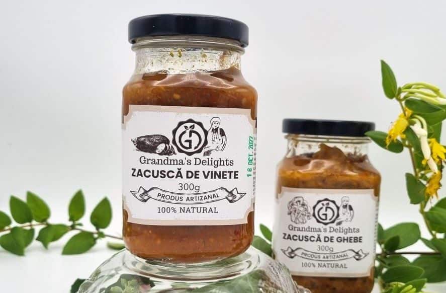 două borcane de zacuscă de la producători sunt asezate intr-un decor cu frunze și un bol de sticlă