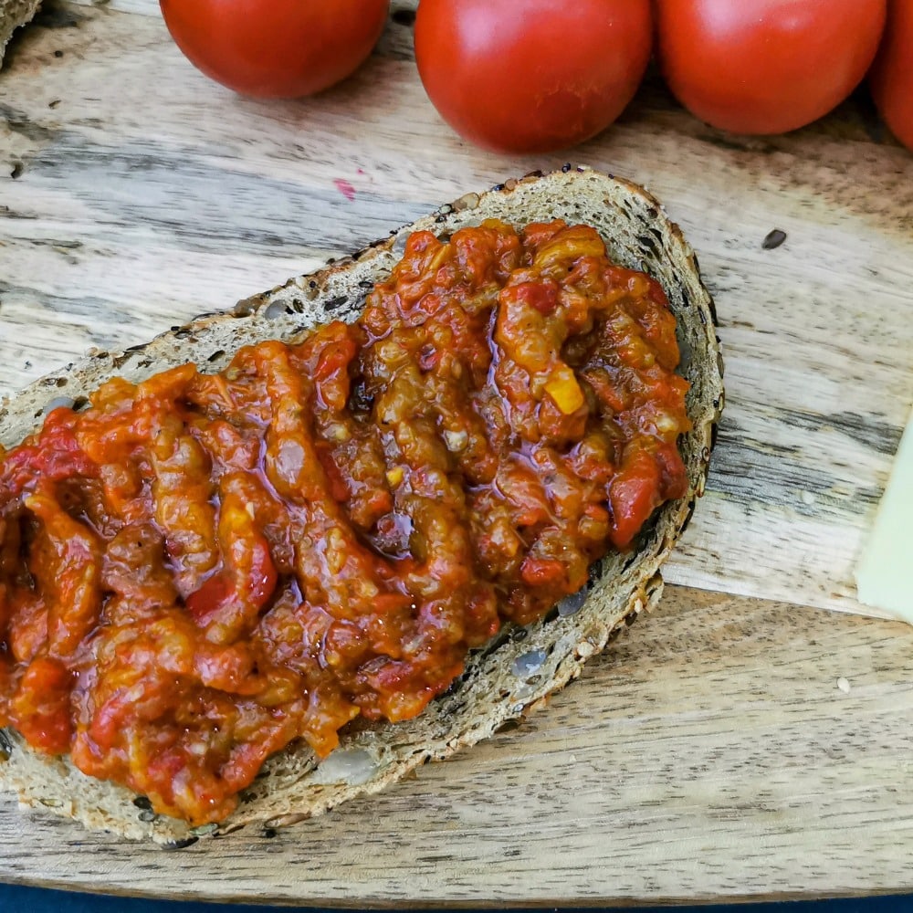 o felie de pâine tărănească cu zacuscă de la producători locali pe o masă de lemn, alături de câteva roșii coapte