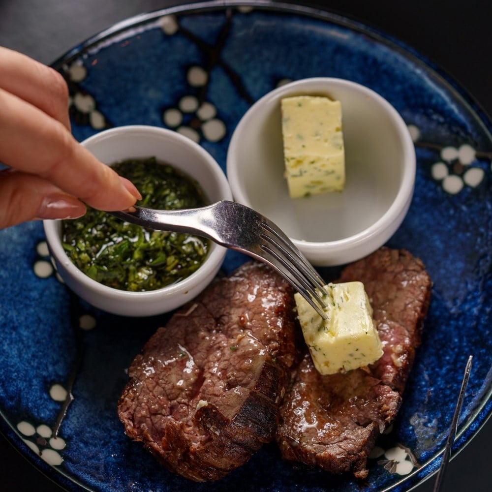 2 felii de steak de vita servite cu unt si sos verde, in farfurie albastra