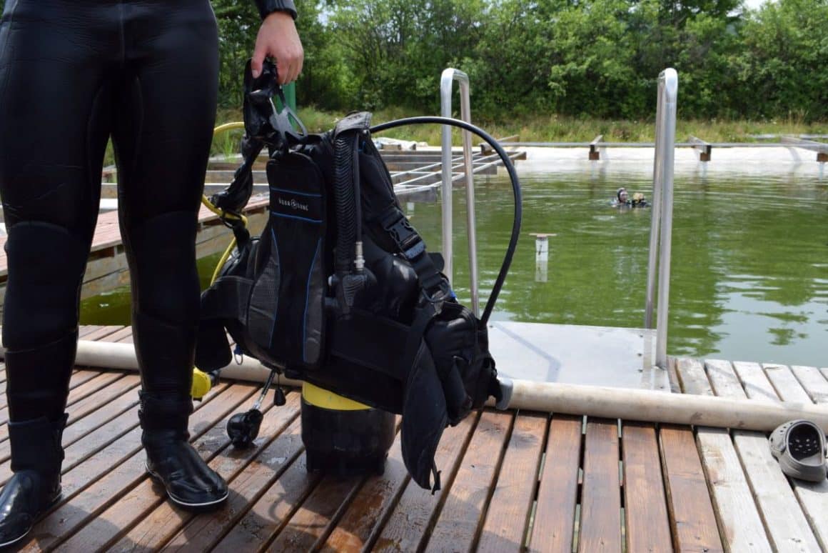 un barbat imbracat in costum de neopren tine in mana, asezat pe ponton, un costum de scafandru