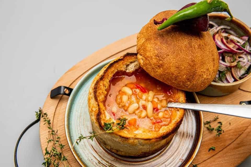 o porție de ciorba de fasole servită în pâine este asezata pe o farfurie modernă, cu o crenguță de cimbru