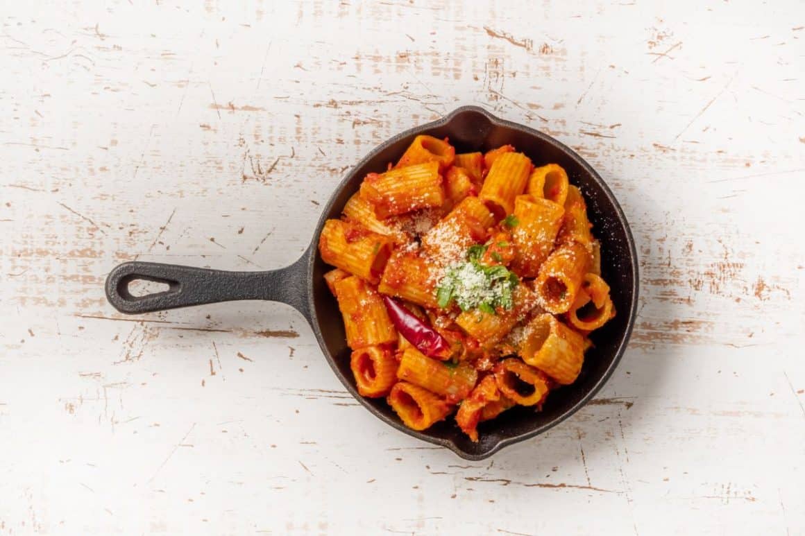 o portie de paste cu Sos Amatriciana, servite intr-o tigaie de fontă, pusă pe o masă de lemn vopsită alb