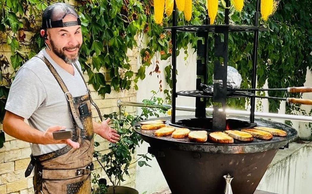 un bucătar pregătește o experiență culinara on demand cu bruschete pe un grătar circular, deasupracăruia sunt atârnați câțiva știuleți de porumb. Experiențe gastronomice inedite. CHef privat