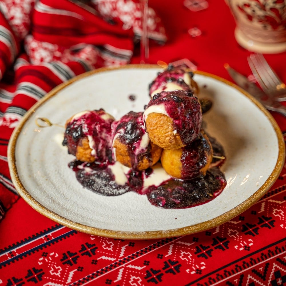 close up cu o farfurie plina cu bilute de papanasi cu smantana si dulceata, asezate pe un stergar rosu cu motive traditionale, de la Casa Românească restaurant, unde mâncăm papanași buni în București