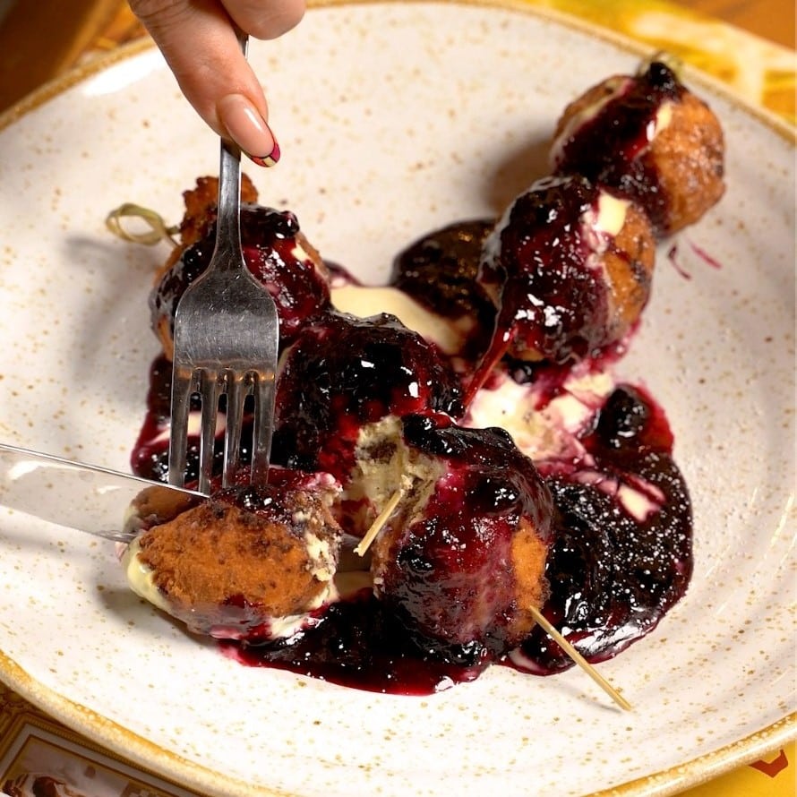 close up cu o farfurie plina cu bilute de papanasi cu smantana si dulceata, din care cineva ia cu furculita si cutit