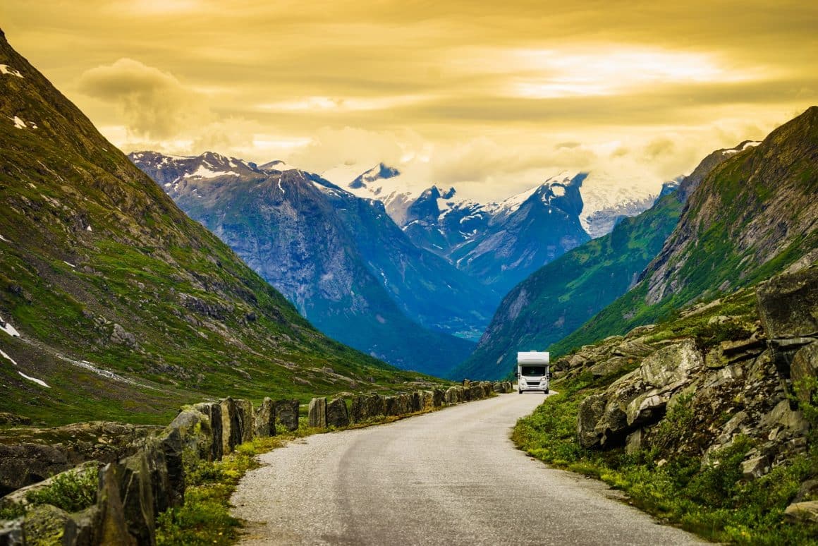 Autorulota fotografiata pe un drum de munte, cu muntii cu varfuri inzapezite, fotografiti la apus. Vacanță cu rulota