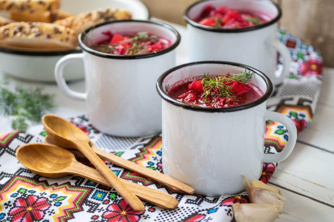 trei căni emailate de bors rusesc cu sfeclă sunt asezate pe un stergar cu motive traditionale și trei linguri de lemn. Povestea borșului.