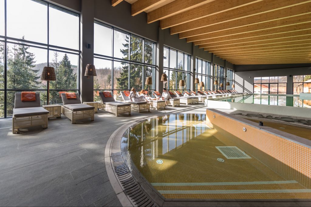 mai multe chez-longuri sunt asezate pe marginea unui spa, iar pe geamurile mari din fundal se vede pădurea. Balvanyos Resort, Grand Santerra Spa. Locații spa din România