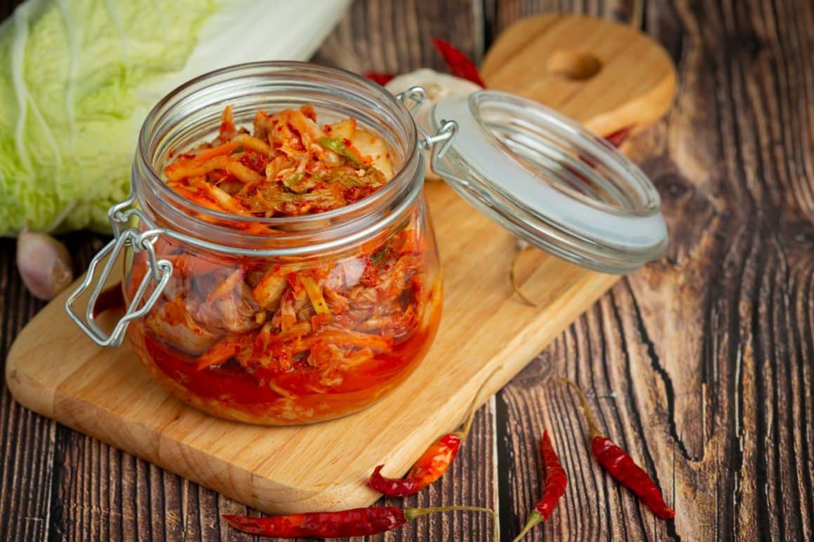 un borcan de kimchi  traditional coreean asezat pe un tocător de lemn și câțiva chilli roșii langa
