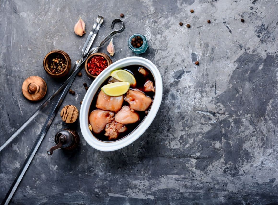 mai multe bucăți de carne de pui sut puse intr-un vas cu marinade pentru carne alaturi de două sferturi de lămâie și alte condimente pe lângă