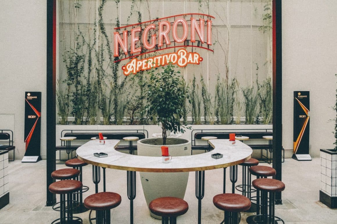 curtea interioara de la Negroni Apertivo Bar București, cu o masa circulara si scaunde bar, iar in fundal silga luminoasa a localului