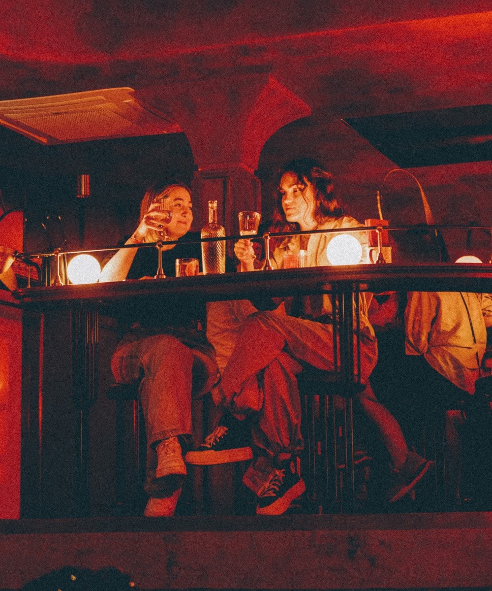 doua femei beau un pahar de cocktail intr-un bar cu lumina difuza, rosiatica