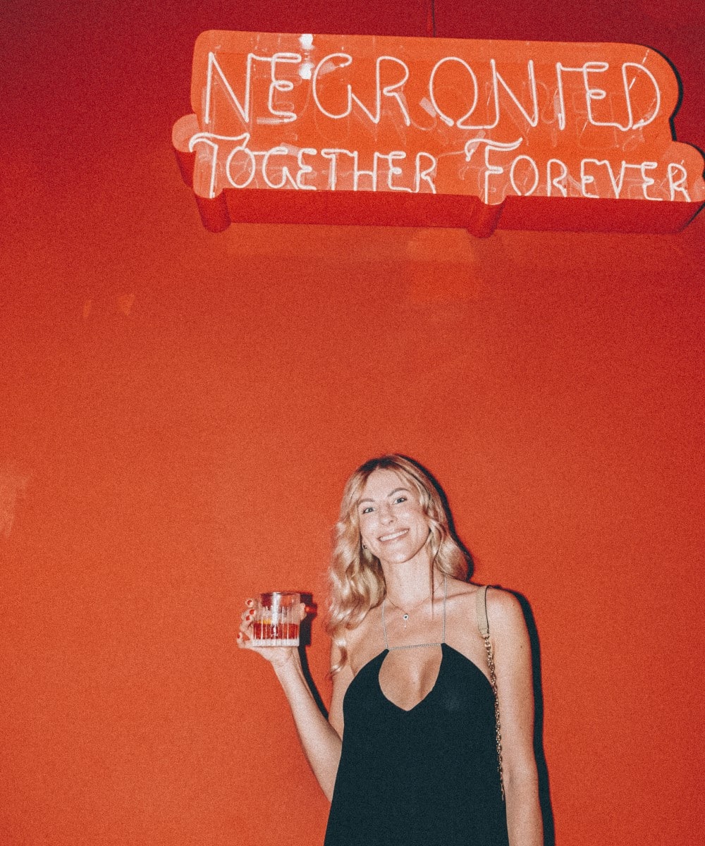 Femeie blonda in rochie neagra, fotografiata in dreptul unui perete rosu cu sigla luminoas aNegroni