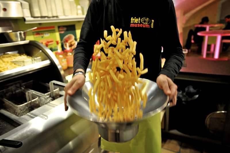 un barbat pune sare peste o portie de cartofi prăjiți și îi amestecă într-un bol special. De mâncat în Bruges