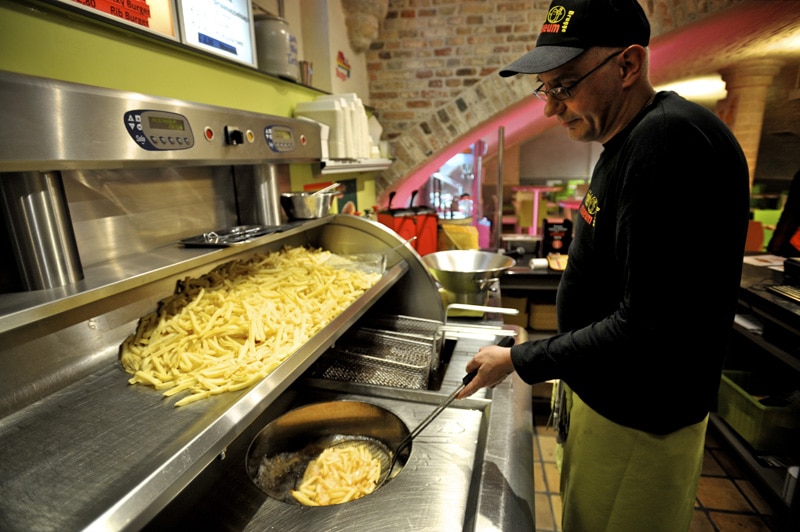 un barbat cu ochelari si sapcă scoate din uneiul încins al unei friteoze o portie de cartofi prăjiți , un fel tipic de mâncare în bruges