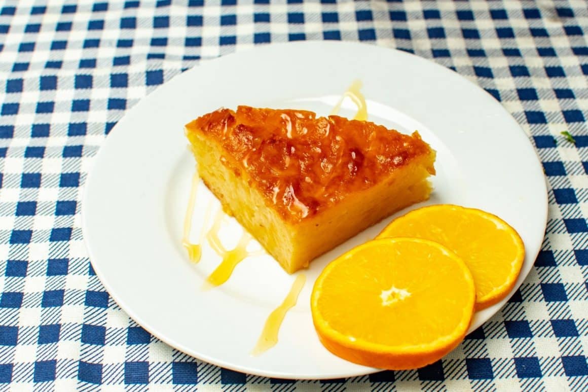 felie mare de plăcintă greceasca cu portocale, pe o farfurie alba alaturi d e2 felii rotunde de portocale