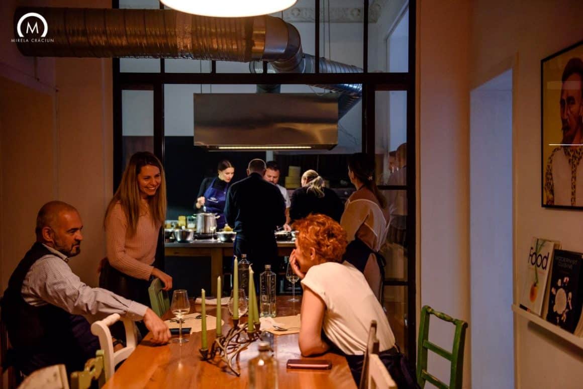 mai ulți adulți socializează în jurul unei mese, în timp ce alții sunt în bucătărie, lângă o altă masă