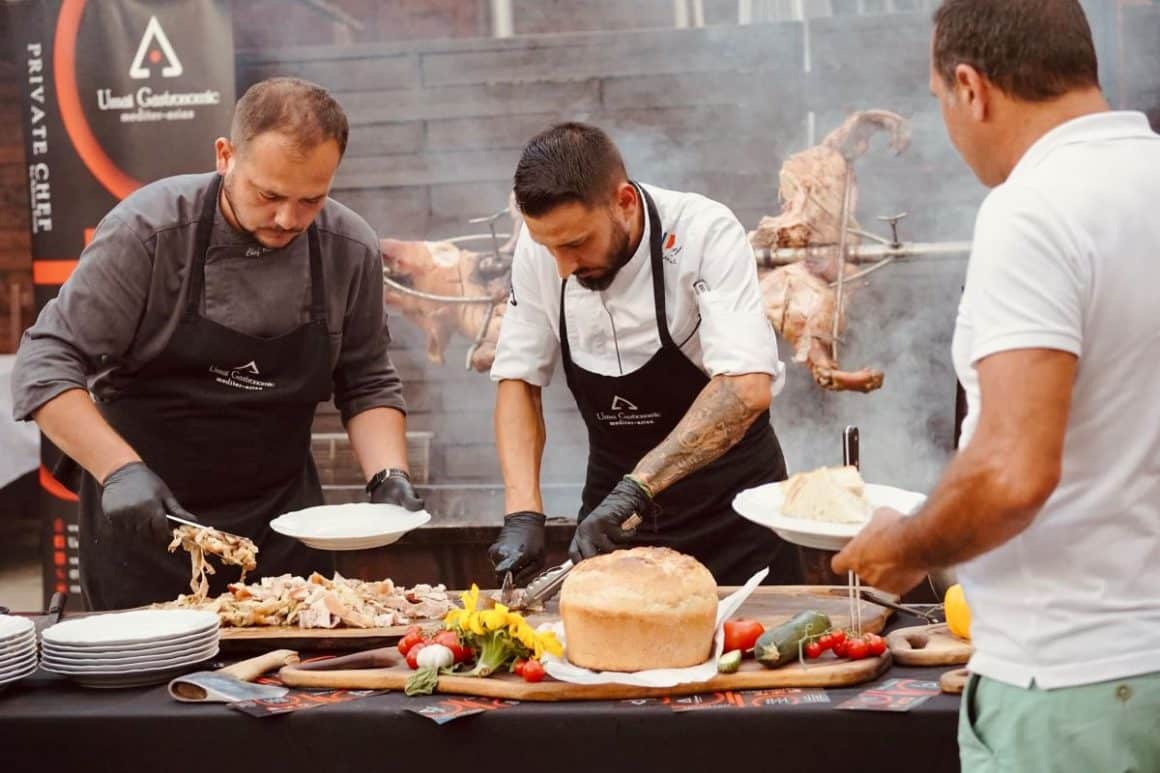 mai mulți bucătari pregătesc un platou de mancare în timp ce în spatele lor se prepară un proțap