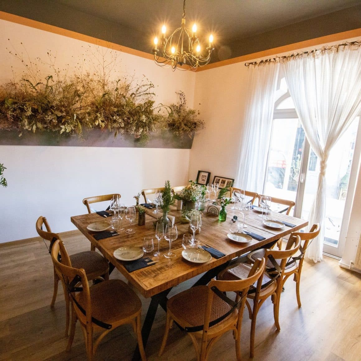 masa mare intr-o camera alba, cu lemn si decoratiuni din plante, un candelabru si ferestre mari, la Bacania Veche Romana, unul din restaurante cozy București