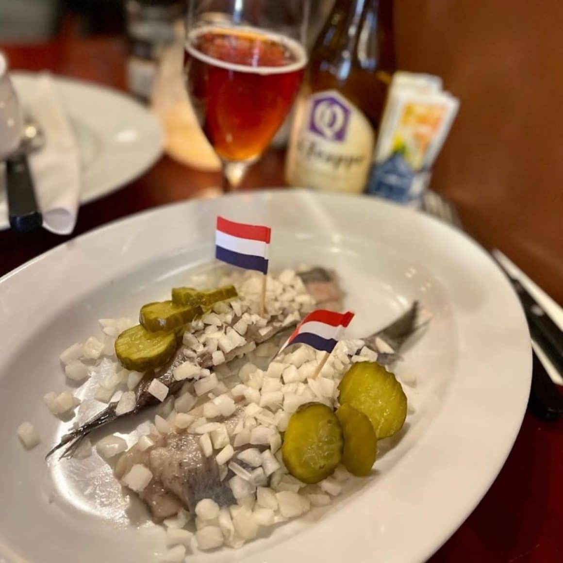 o portie de hering marinat cu ceapă și castraveți murați, intr-o farfurie albă, cu două stegulețe olandeze