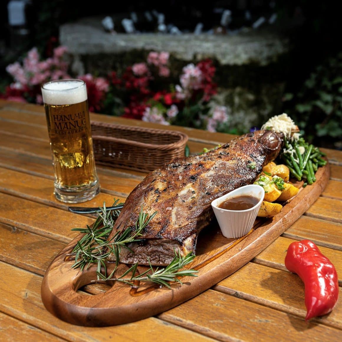 o portie generoasă de carne la grătar, servită peun platou tradițional de lemn, cu crenguțe de rozmarin, cartofi, un sos și o halbă de bere