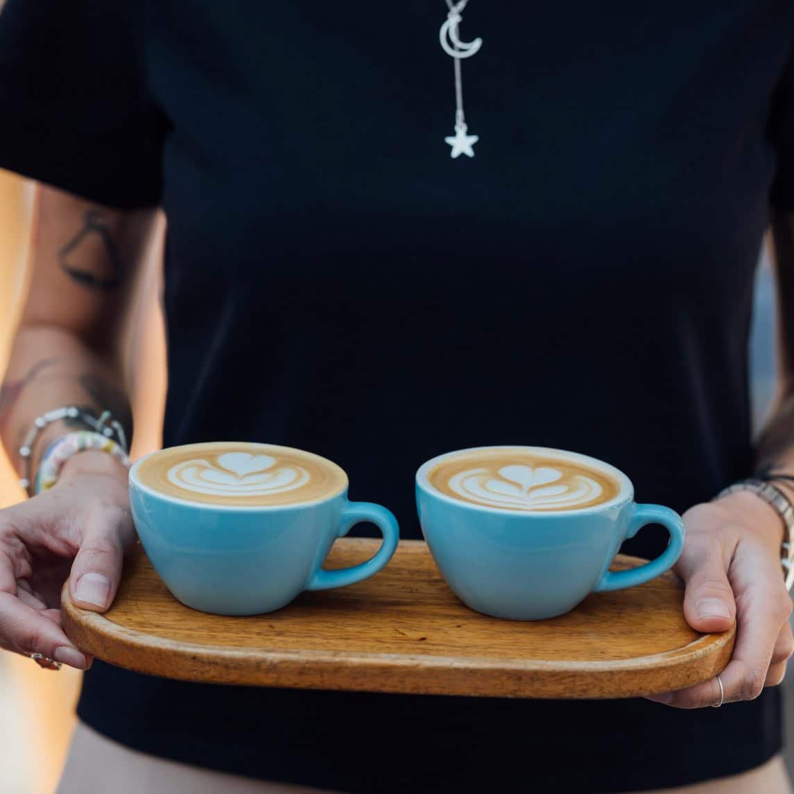 un bărbat tânăr cu tatuaje duce pe o tavă de lemn două cești de cafea