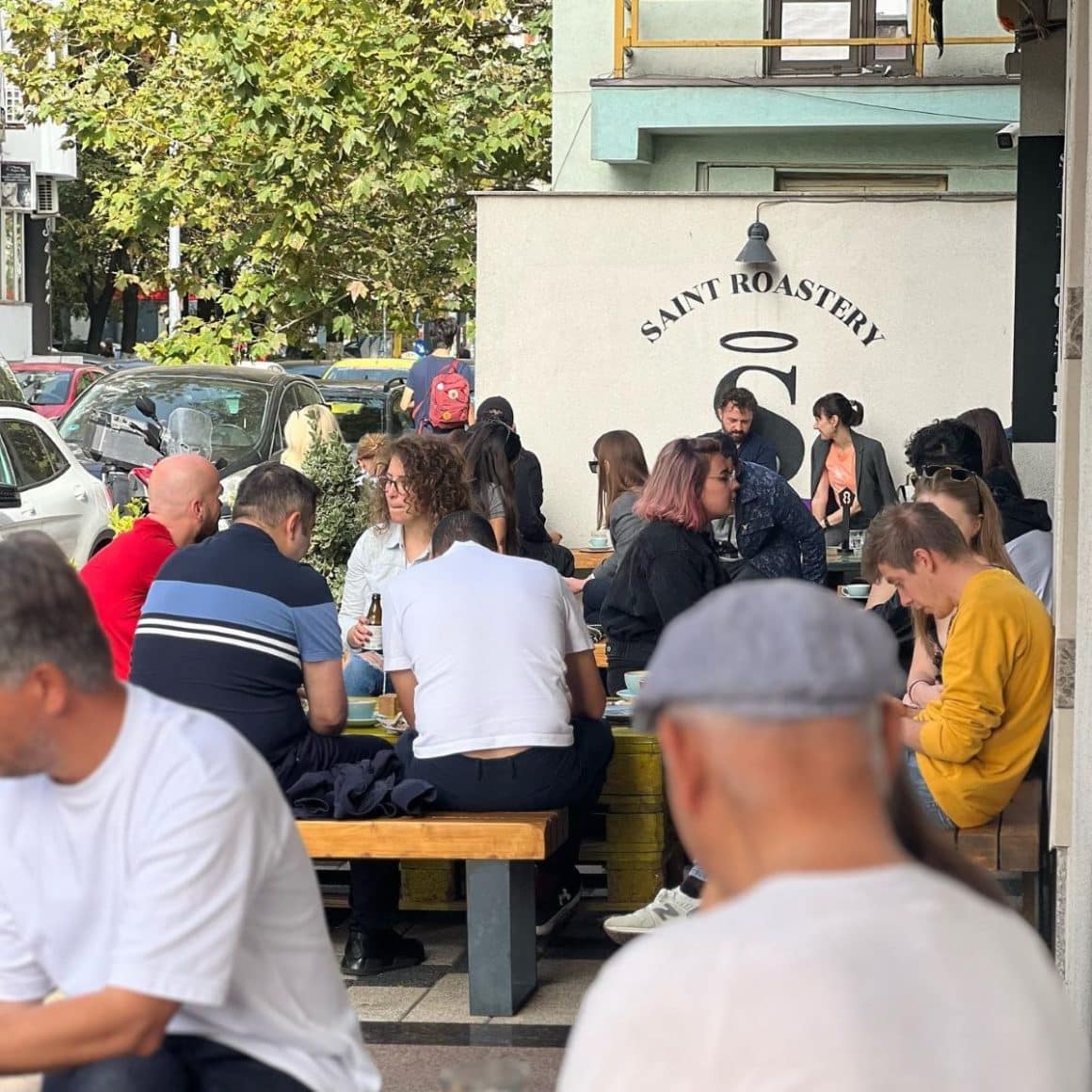 mai multi tineri socializează în fata unei cafenele micute din bucuresti, stând la mese de lemn