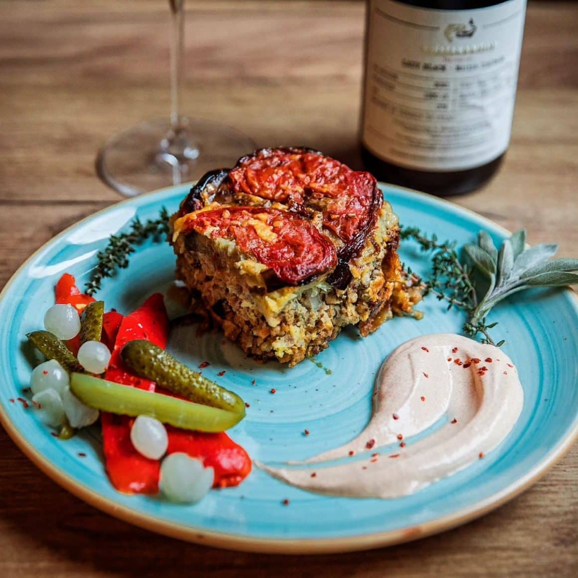 o porție de musaca tradițională din  bucătăria românească, alături de murături și un sos, pe o farfurie modernă