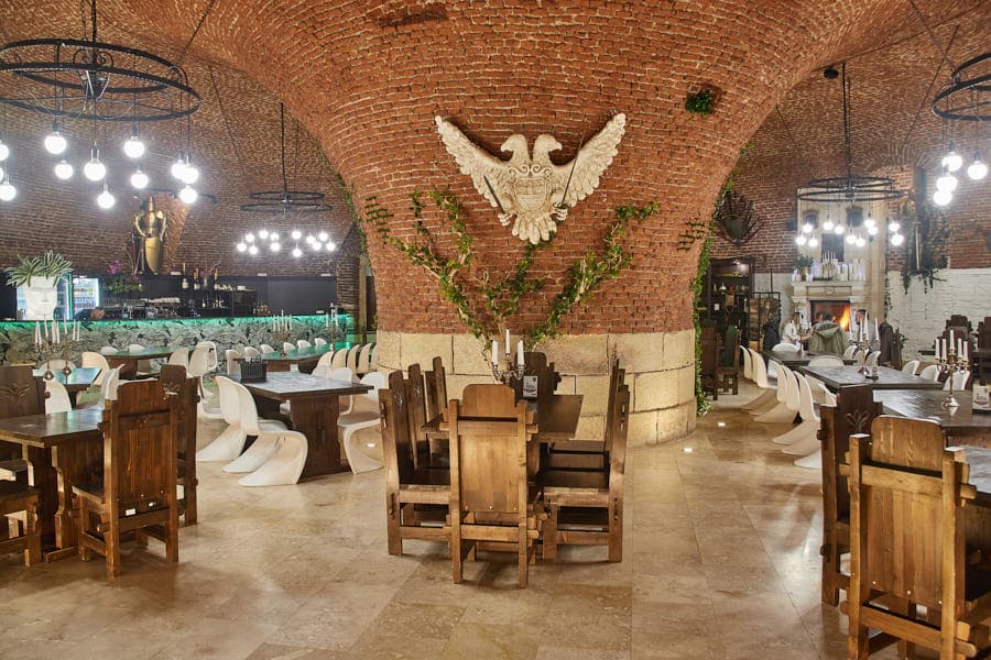 mai multe mese și scaune de lemn sunt asezate intr-un restaurant din alba iulia cu o coloană masivă de cărămidă roșie