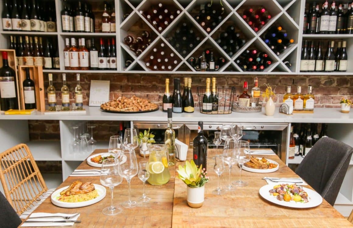 masa de 4 persoane cu farfurii cu mancare si vin in dreptul unor rafturi pline cu sticle de vin la Bacania Veche Romana, unul din restaurante cozy București