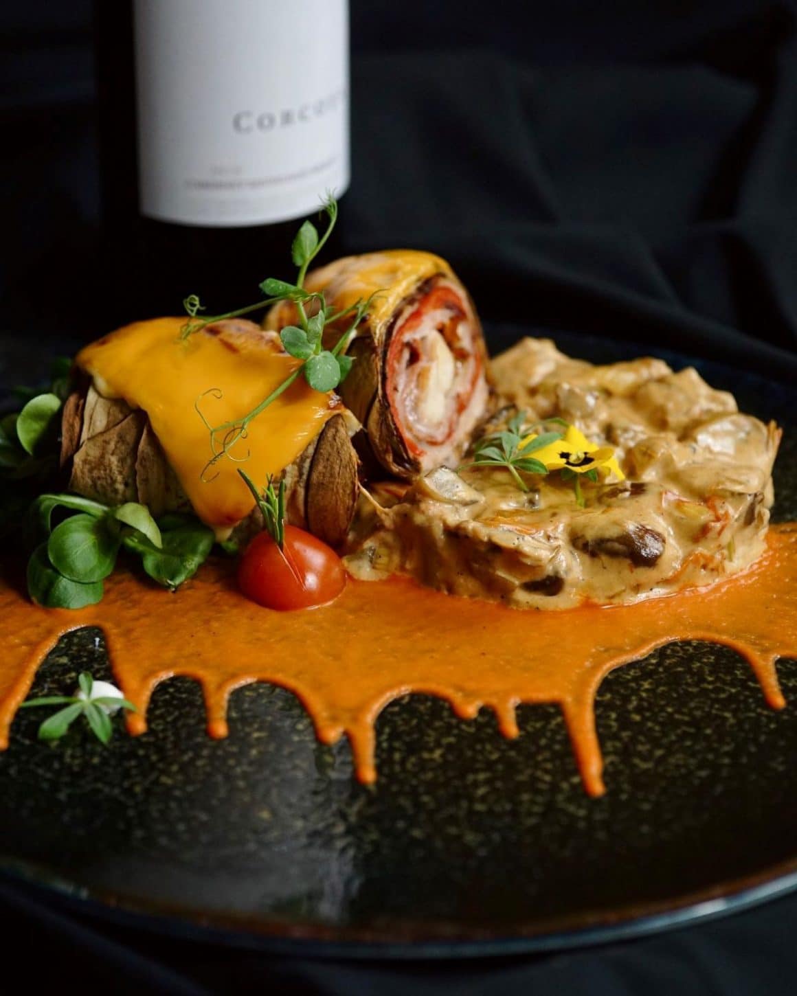 o porție de mâncare cu sos și micro-planteeste servită pe o farfurie neagră, într-un restaurant din alba iulia, alături de o sticlă de vin