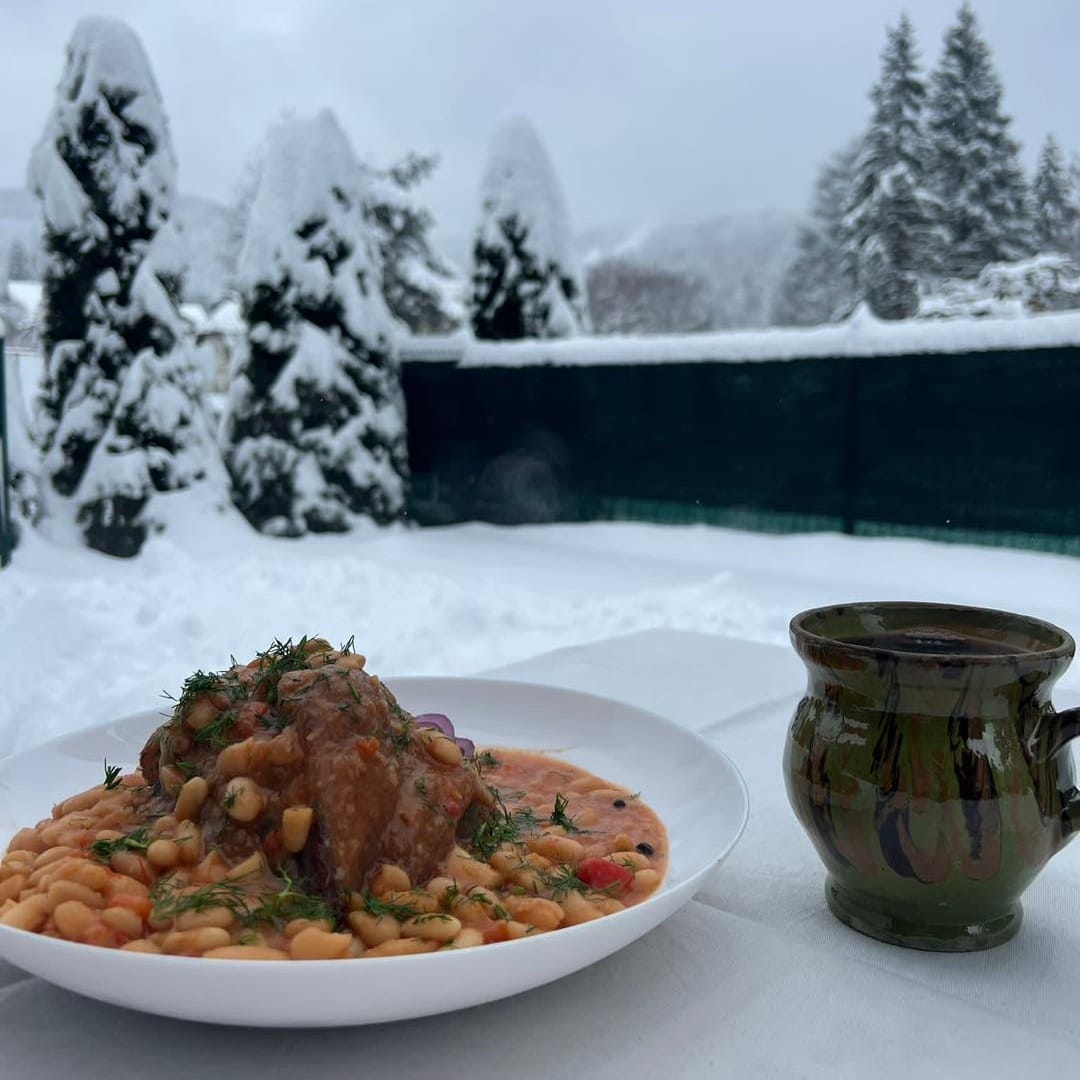 farfurie cu iahnie de fasole si cioan, langa o ceasca de lut cu vin, pe o masa afara, in zapada